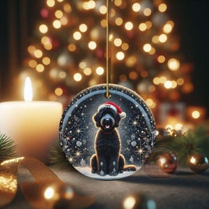 May include: A round Christmas ornament featuring a black dog wearing a Santa hat and red collar, set against a snowy winter scene. The ornament is hanging with a gold string, with a lit candle and Christmas tree lights in the background.