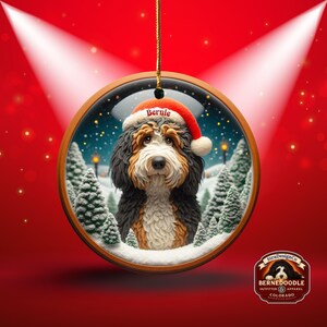 Op de afbeelding: Een ronde kerstversiering met een Bernedoodle-hond met een kerstmuts met de naam "Bernie". De versiering heeft een winterse achtergrond met besneeuwde bomen en een besneeuwde grond. De versiering hangt aan een gouden touwtje.