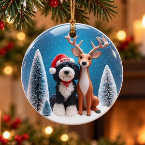 May include: A round ceramic Christmas ornament featuring a black and white dog wearing a red Santa hat with the name Bernie printed on it, sitting next to a brown deer in a snowy forest with evergreen trees under a blue sky with falling snow.