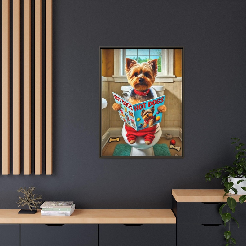May include: A framed print featuring a Yorkshire Terrier sitting on a toilet, reading a magazine titled "HOT DOGS." The dog is wearing red pants and a red collar. The background shows a bathroom scene with a window.