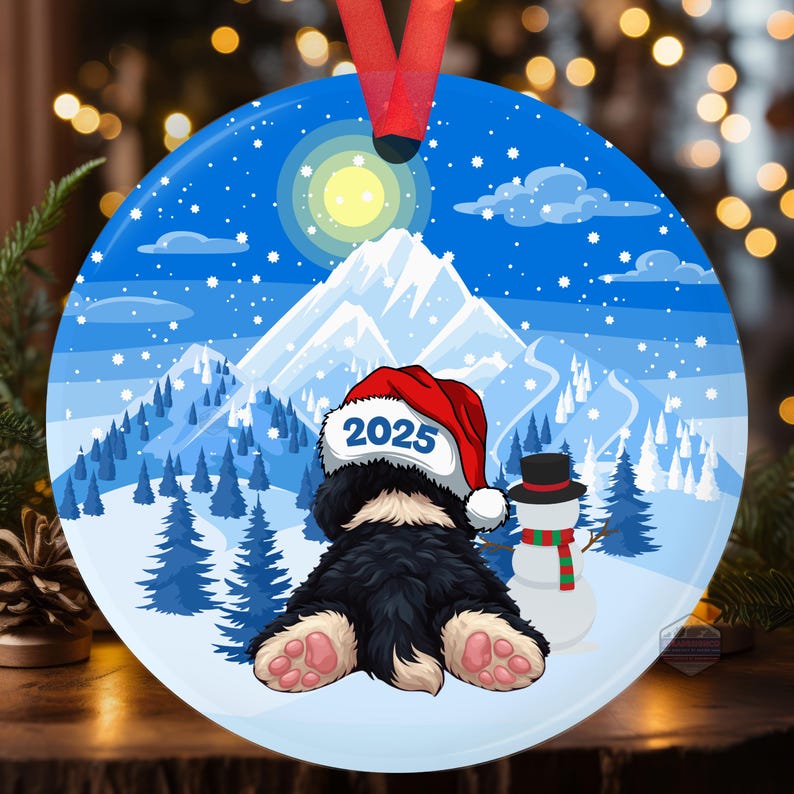 May include: Round Christmas ornament featuring a black and white dog wearing a Santa hat with "2025" on it. A snowman with a red and green scarf stands nearby. The background shows a snowy mountain scene under a blue sky.