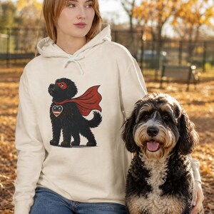 Pu&ograve; includere: Una felpa con cappuccio color crema che presenta la grafica di un cane nero che indossa un mantello da supereroe rosso, una maschera rossa e un emblema a forma di cuore sul petto con un motivo a stelle blu, bianco e rosso.