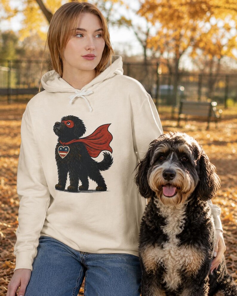 Pu&ograve; includere: Una felpa con cappuccio color crema che presenta la grafica di un cane nero che indossa un mantello da supereroe rosso, una maschera rossa e un emblema a forma di cuore sul petto con un motivo a stelle blu, bianco e rosso.