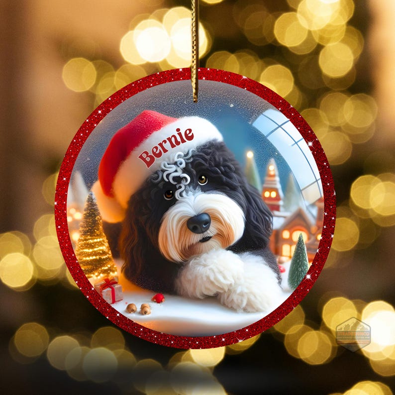K&ouml;nnte beinhalten: Runder Weihnachtsschmuck mit einem schwarz-wei&szlig;en Hund, der einen Weihnachtsmannhut mit dem Namen "Bernie" tr&auml;gt. Der Schmuck hat einen roten Glitzerrahmen und eine goldene Schnur zum Aufh&auml;ngen. Der Hintergrund zeigt eine Winterlandschaft mit Schnee, B&auml;umen und H&auml;usern.