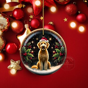 May include: A round Christmas ornament featuring a golden doodle dog wearing a Santa hat with the name "Goldie". The ornament is surrounded by holly and berries, set against a snowy background. The ornament hangs from a gold string.