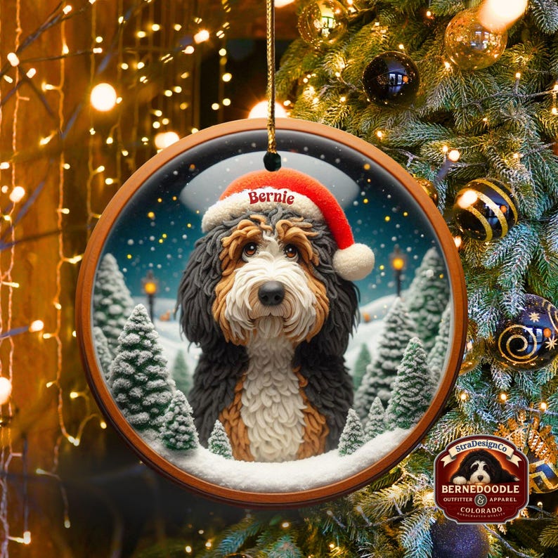Op de afbeelding: Ronde houten kerstversiering met een Bernedoodle-hond met een kerstmuts met de naam "Bernie". De hond staat tegen een besneeuwde wintersc&egrave;ne met dennenbomen. De versiering hangt aan een gouden touwtje.