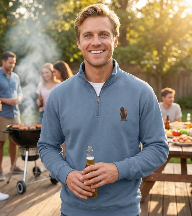 Pu&ograve; includere: Una felpa blu chiaro con colletto con zip e un piccolo disegno di cane ricamato. La persona tiene in mano una bottiglia di birra. Altre persone sono sullo sfondo vicino a un barbecue e a un tavolo da picnic.