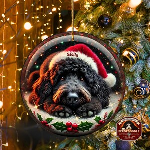 May include: Round Christmas ornament featuring a black dog wearing a Santa hat. The dog's name, "Stella," is printed on the hat. The ornament has a festive design with holly and berries, and is hanging from a gold string. The background is a Christmas tree.
