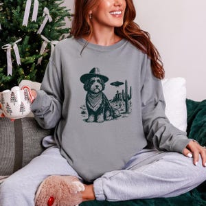 May include: A gray long-sleeve sweatshirt featuring a graphic of a dog wearing a cowboy hat and bandana, set against a desert landscape with a UFO. The dog is the central focus of the design.