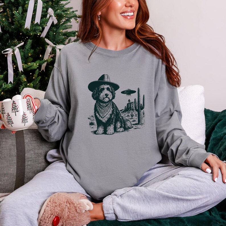 May include: A gray long-sleeve sweatshirt featuring a graphic of a dog wearing a cowboy hat and bandana, set against a desert landscape with a UFO. The dog is the central focus of the design.