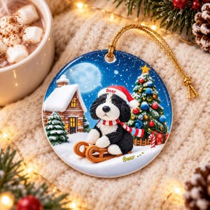A ceramic ornament featuring a black and white dog wearing a Santa hat and scarf, sitting on a sled and looking at the camera, with a snowy village scene and a Christmas tree in the background.