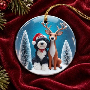 May include: A round ceramic Christmas ornament featuring a black and white dog wearing a red Santa hat with the name Bernie printed on it, sitting next to a brown deer in a snowy forest with pine trees under a blue sky with a gold string.