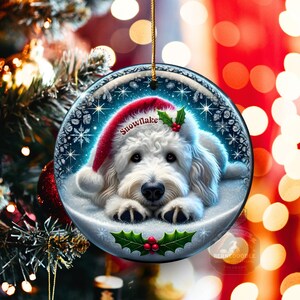 May include: A round Christmas ornament featuring a white dog wearing a red Santa hat with the word "Snowflake". The ornament has a snowy background with snowflakes and holly accents. A gold string is attached for hanging.