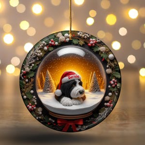 May include: A round Christmas ornament featuring a snow globe design with a black and white dog wearing a Santa hat that says "Bernie". The ornament is surrounded by a wreath of holly and berries, with a red bow at the bottom.