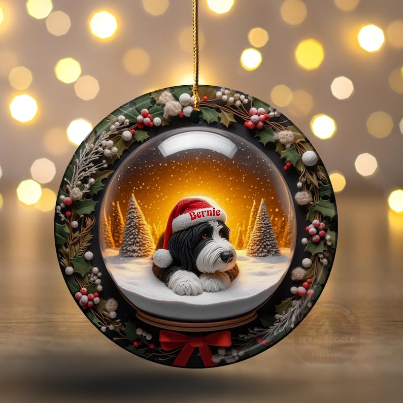 May include: A round Christmas ornament featuring a snow globe design with a black and white dog wearing a Santa hat that says "Bernie". The ornament is surrounded by a wreath of holly and berries, with a red bow at the bottom.