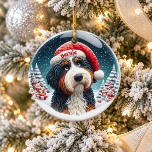 May include: A round ceramic Christmas ornament featuring a Bernese Mountain Dog wearing a red Santa hat with the name Bernie printed on the brim. The dog is set against a snowy winter landscape with pine trees and small gift boxes.
