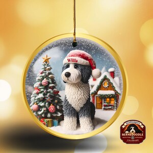 May include: A circular Christmas decoration featuring a Bernedoodle dog wearing a Santa hat with the name "Willis". The ornament depicts a snowy winter scene with a Christmas tree and a house. The decoration is trimmed with gold.