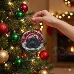 May include: A round Christmas ornament featuring a black dog wearing a Santa hat with the name "Teddi". The ornament is hanging on a Christmas tree with red and green ornaments and lights. The ornament has a gold string.