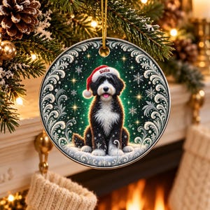 May include: A round ceramic Christmas ornament featuring a black and white Bernedoodle dog wearing a red Santa hat with the name Stella printed on it. The dog sits against a dark green background with white snowflakes and ornate silver borders.