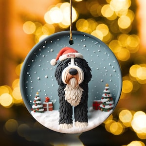 May include: A round Christmas ornament featuring a black and white dog wearing a Santa hat with the name "Willy". The ornament has a snowy scene with Christmas trees and presents. A gold string is attached for hanging.