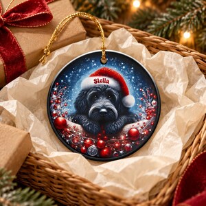 May include: A round ceramic Christmas ornament featuring a black dog wearing a red and white Santa hat with the name Stella printed in red. The background shows a snowy night scene with red berries and ornaments hanging from a gold string.