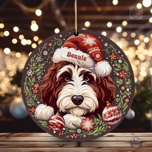 Op de afbeelding: Ronde kerstversiering met een hond met een kerstmuts en de naam "Bennie". De versiering heeft een bruine achtergrond met een decoratieve bloemenrand en twee rood-witte ballen. Een gouden draad is bevestigd om op te hangen.