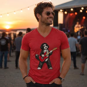 May include: A red V-neck t-shirt featuring a graphic illustration of a black and white dog playing a red electric guitar. The shirt has short sleeves and a standard fit, showcasing a detailed print of the canine musician centered on the chest.