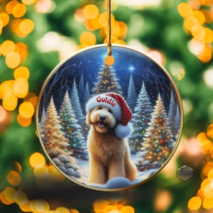May include: A round Christmas ornament featuring a golden doodle wearing a Santa hat with the name "Goldie" on it. The ornament has a winter scene with snow-covered trees and a starry sky. The ornament is hanging from a gold string.