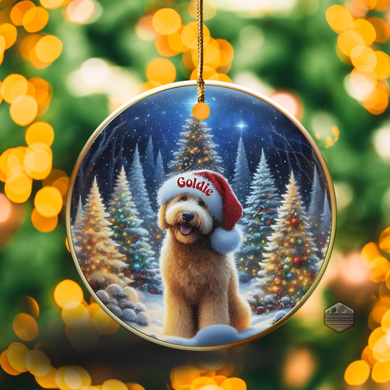 May include: A round Christmas ornament featuring a golden doodle wearing a Santa hat with the name "Goldie" on it. The ornament has a winter scene with snow-covered trees and a starry sky. The ornament is hanging from a gold string.