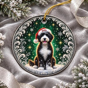 May include: A round ceramic Christmas ornament featuring a black and white Bernedoodle dog wearing a red Santa hat with the name Stella printed on the brim. The background is a dark green gradient with white snowflakes and gold star accents.