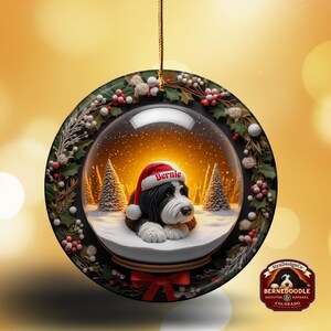 May include: A round Christmas ornament featuring a Bernedoodle dog inside a snow globe. The dog wears a Santa hat with the name "Bernie". The ornament is decorated with holly and berries, and has a red bow at the bottom.