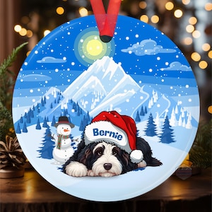 May include: Round Christmas ornament with a winter scene featuring a black and white dog wearing a Santa hat with the name "Bernie". The background includes a snowman, snow-covered mountains, and pine trees under a blue sky.