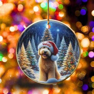 May include: A round Christmas ornament featuring a golden retriever wearing a Santa hat with the name "Goldie" on it. The background shows a snowy forest with Christmas trees. The ornament has a gold trim and a gold string for hanging.