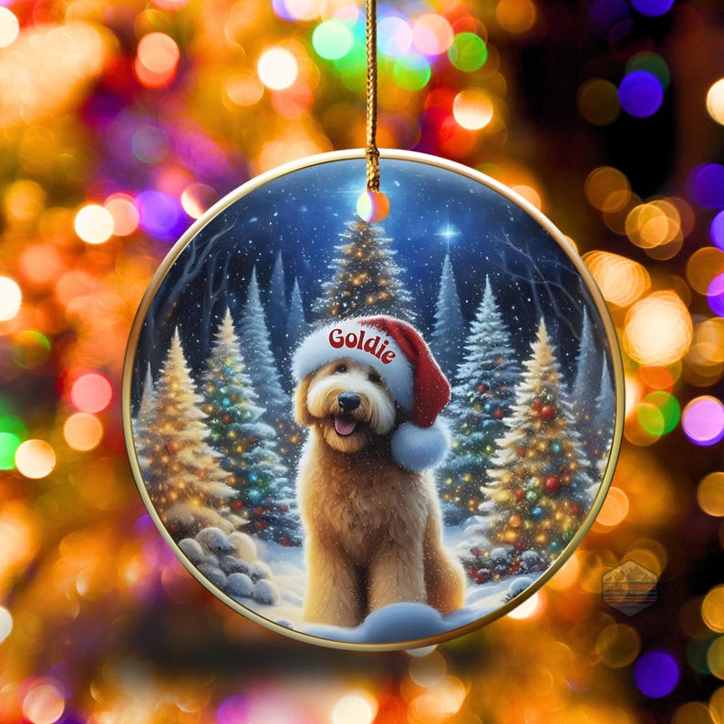 May include: A round Christmas ornament featuring a golden retriever wearing a Santa hat with the name "Goldie" on it. The background shows a snowy forest with Christmas trees. The ornament has a gold trim and a gold string for hanging.