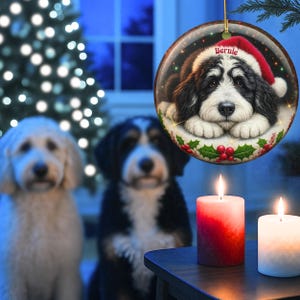 May include: A round Christmas ornament featuring a black and white dog wearing a Santa hat, with the name "Bernie" on it. The ornament is hanging in front of a Christmas tree and two lit candles. Two dogs are in the background.