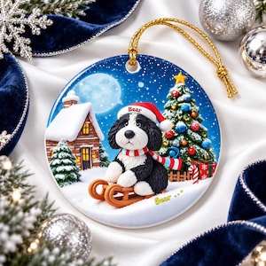 A ceramic ornament featuring a black and white dog wearing a Santa hat, sitting on a sled and looking at the camera, with a snowy village scene and a Christmas tree in the background.