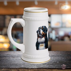 May include: White ceramic beer stein with a handle and a gold rim. The stein features a detailed illustration of a Bernese Mountain Dog with a tag that says "Walter". The background is blurred, suggesting a cafe setting.