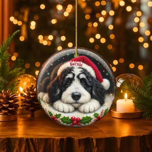 May include: Round Christmas ornament featuring a black and white dog wearing a Santa hat. The dog's name, "Bernie", is printed on the hat. The ornament is decorated with holly and red berries.