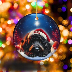 May include: Round Christmas ornament featuring a black dog wearing a Santa hat with the name "Stella" on it. The background is a starry night scene with a bright star and red accents. A gold string is attached for hanging.