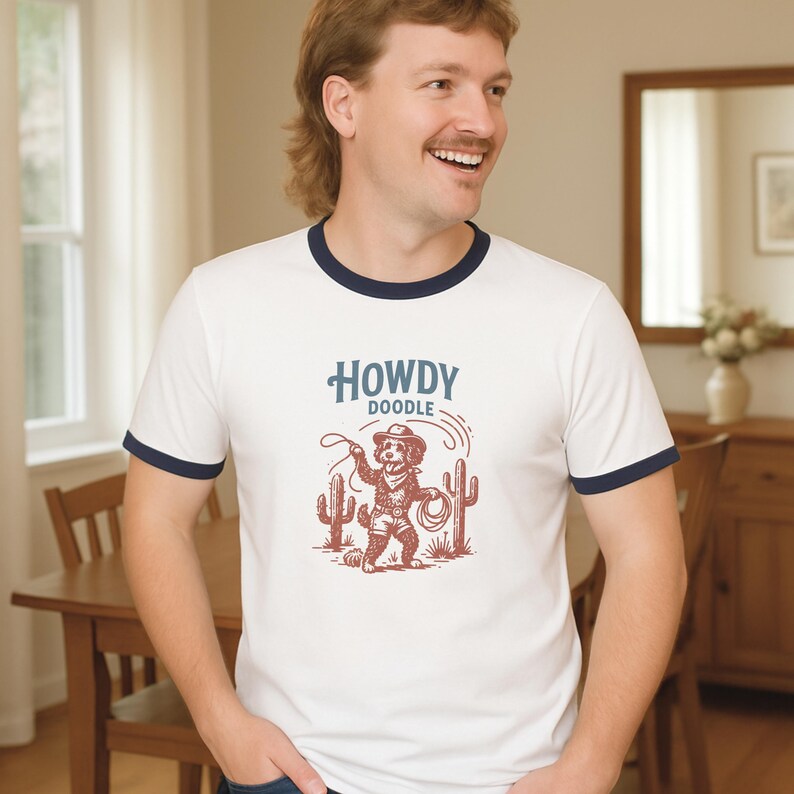 May include: White ringer t-shirt with navy blue trim featuring a cartoon cowboy dog graphic with the text "HOWDY DOODLE". The dog is holding a lasso with cacti on either side. The shirt is being worn by a person.