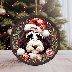 May include: A round Christmas ornament featuring a black and white dog wearing a red Santa hat with the name "Bernie". The ornament is decorated with red and white ornaments and a green wreath.