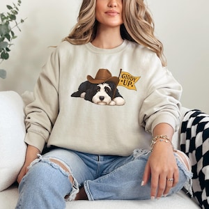 Puede incluir: Sudadera beige con un gr&aacute;fico de un perro blanco y negro con un sombrero de vaquero marr&oacute;n. El perro sostiene una bandera amarilla que dice "Giddy Up". La sudadera tiene mangas largas y cuello redondo.