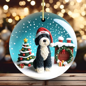 May include: A round Christmas ornament featuring a black and white dog wearing a Santa hat with the name "Mabel". The ornament has a winter scene with a Christmas tree, snow, and a decorated chimney.