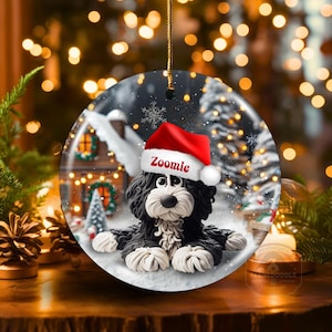 May include: A round Christmas ornament featuring a black and white dog wearing a red Santa hat with the name "Zoomie". The ornament has a winter scene background with a house and snow. A gold string is attached for hanging.