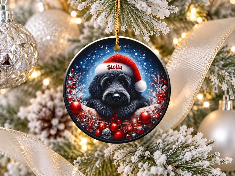 May include: A round ceramic Christmas ornament featuring a black dog wearing a red Santa hat with the name Stella printed on the brim. The dog is surrounded by red berries and ornaments against a dark blue, snowy, starry background.