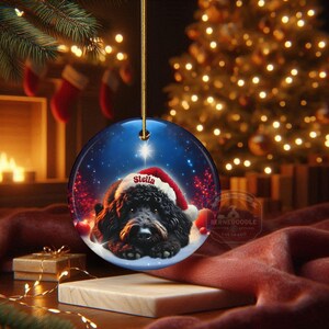 May include: A round Christmas ornament featuring a black dog wearing a Santa hat with the name "Stella" on it. The ornament has a blue background with a star and festive red accents. The ornament is hanging from a gold string.