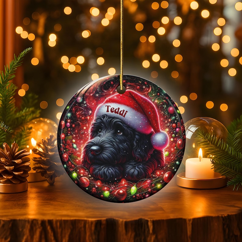 May include: Round Christmas ornament featuring a black dog wearing a Santa hat with the name "Teddi" on it. The ornament has a festive design with red and green accents, and is hanging from a gold string. The background is blurred with bokeh lights.