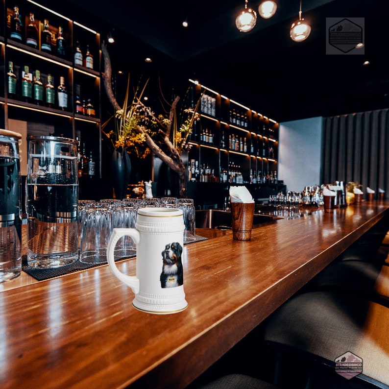 May include: White ceramic beer stein with a handle and a gold rim. The stein features a portrait of a Bernese Mountain Dog. The background shows a bar with bottles of liquor and glasses.
