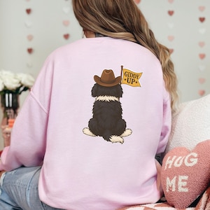 Puede incluir: Sudadera rosa claro con un dise&ntilde;o de un perro blanco y negro con un sombrero de vaquero marr&oacute;n. El perro sostiene una bandera amarilla que dice "Giddy Up". Un coj&iacute;n rosa en forma de coraz&oacute;n con "HUG ME" est&aacute; en la esquina inferior derecha.