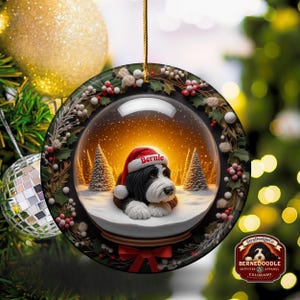 May include: A round Christmas ornament featuring a Bernedoodle dog wearing a Santa hat with the name "Bernie". The ornament is a snow globe design with a snowy scene and pine trees. The ornament is trimmed with holly and berries.
